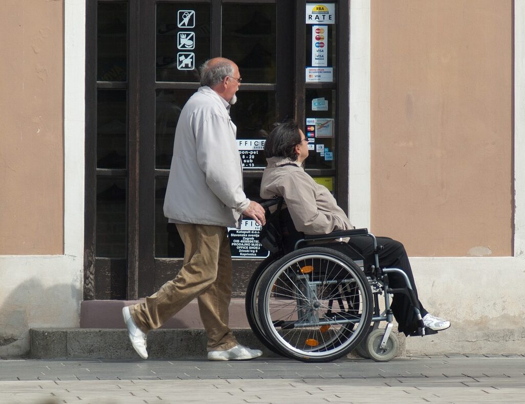 Asistent po pločniku poganja voziček na katerem je invalid